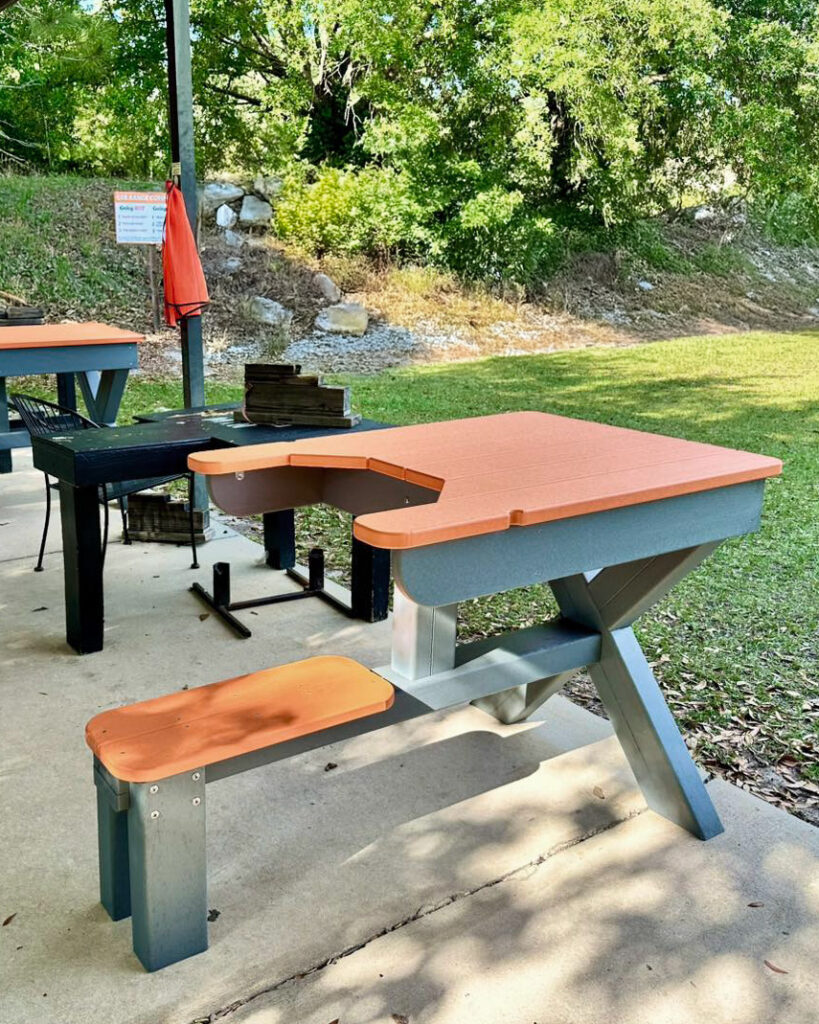 Outdoor shooting bench constructed from durable plastic lumber, featuring an X-shaped support frame, a rectangular tabletop with a shooter cut-out, and an attached seat. Positioned on a concrete pad with grassy surroundings and trees in the background. A second bench and shooting equipment are visible in the distance